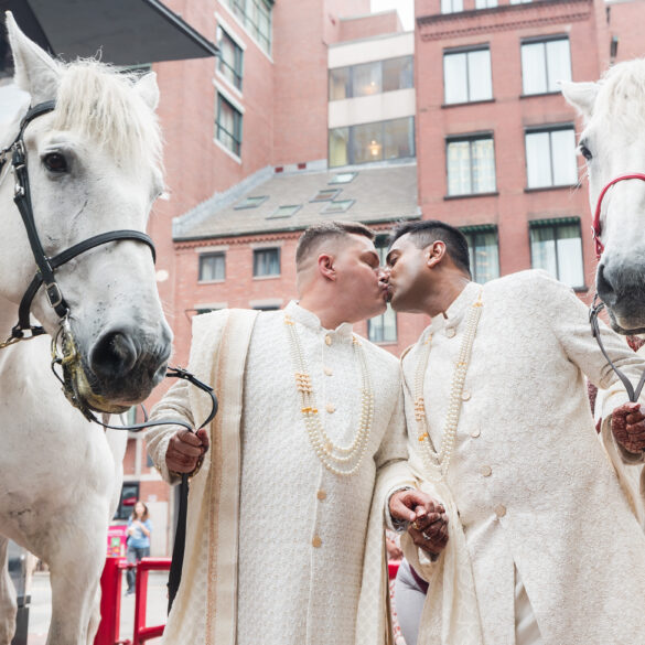 Couple kisses beside two horses at wedding event.